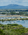 A scenic landscape photograph of a Bowen harbour filled with boats, surrounded by green vegetation and a town, with mountains in the background under a cloudy sky.