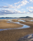 A scenic landscape photograph of a tidal flat or salt marsh with winding shallow water channels reflecting the sky, under a blue sky with scattered white clouds and distant hills.