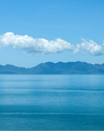 A scenic landscape photograph of a calm blue sea under a bright blue sky with white clouds and distant blue mountains. A small artist signature is visible in the bottom right corner.