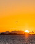 A scenic sunset over water with a mountain silhouette on the horizon, a bright sun near the horizon, and two small helicopters flying across the sky. The sky is a warm golden-orange gradient.