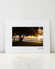 A framed or matted art print leaning against a white wall on a light marble surface. The print shows a sunset seascape with dark trees on the left and golden light over the water and shoreline.