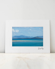 A framed landscape art print featuring a scenic view of a blue lake with mountains in the distance, displayed on a light marble surface against a white wall.