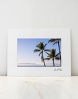 A matted art print featuring a photograph of palm trees against a blue sky, displayed on a light marble surface against a white wall.