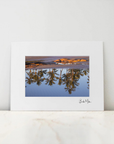 A framed art print with a wide white mat and a central landscape photograph showing a lake reflection, leaning against a white wall on a light marble surface.