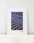 A vertical art print with a wide white border, featuring a photograph of blue and brown wavy dune patterns, leaning against a white wall on a light marble surface.
