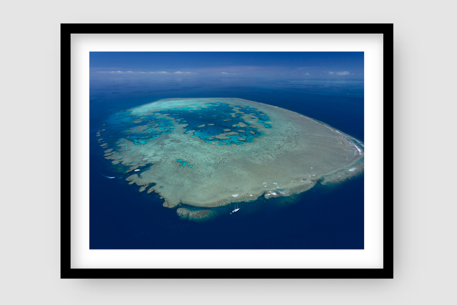 blue waters and sky surrounding large reef island waves breaking with sailing vessel in bottom left