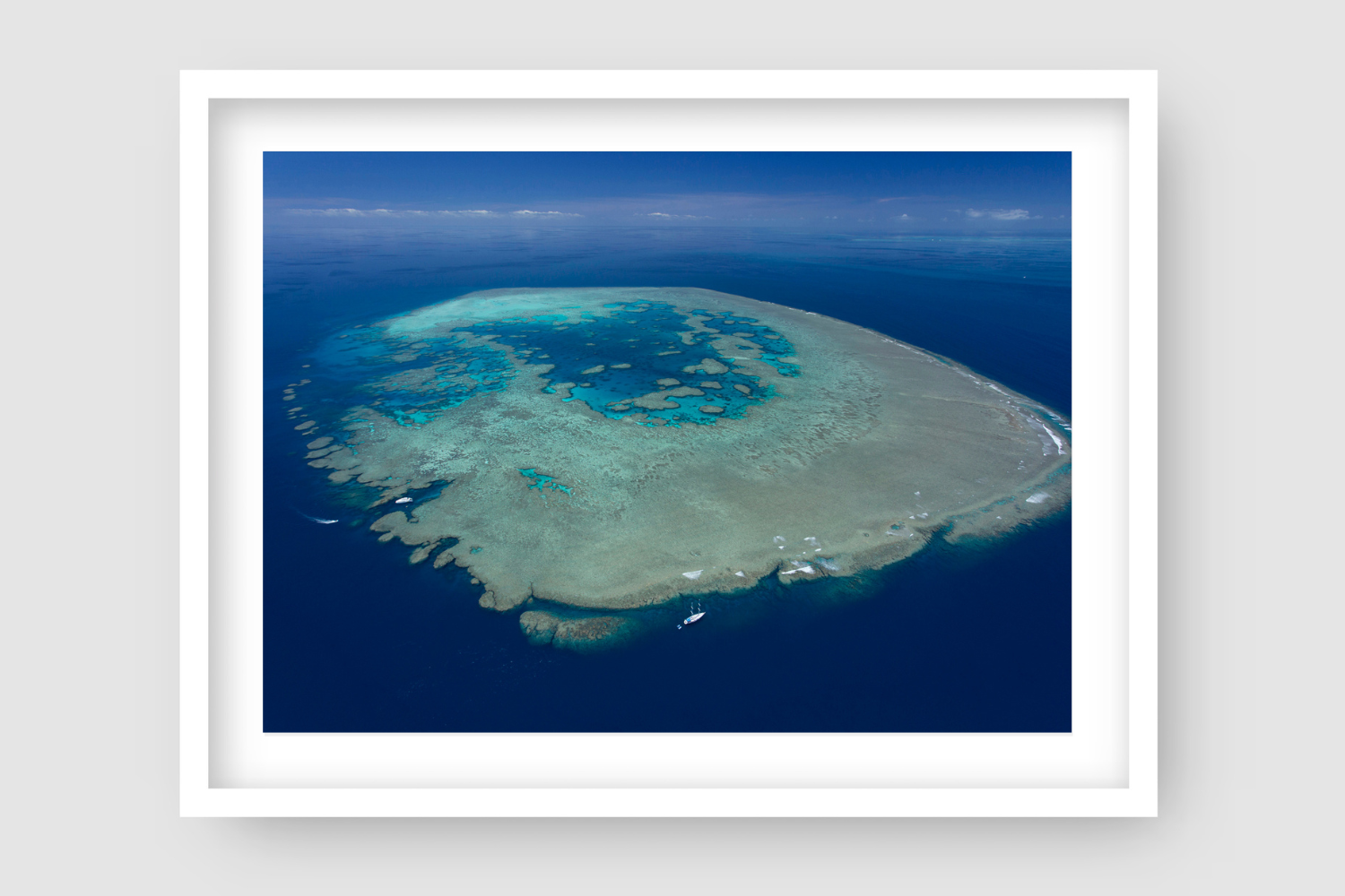 blue waters and sky surrounding large reef island waves breaking with sailing vessel in bottom left