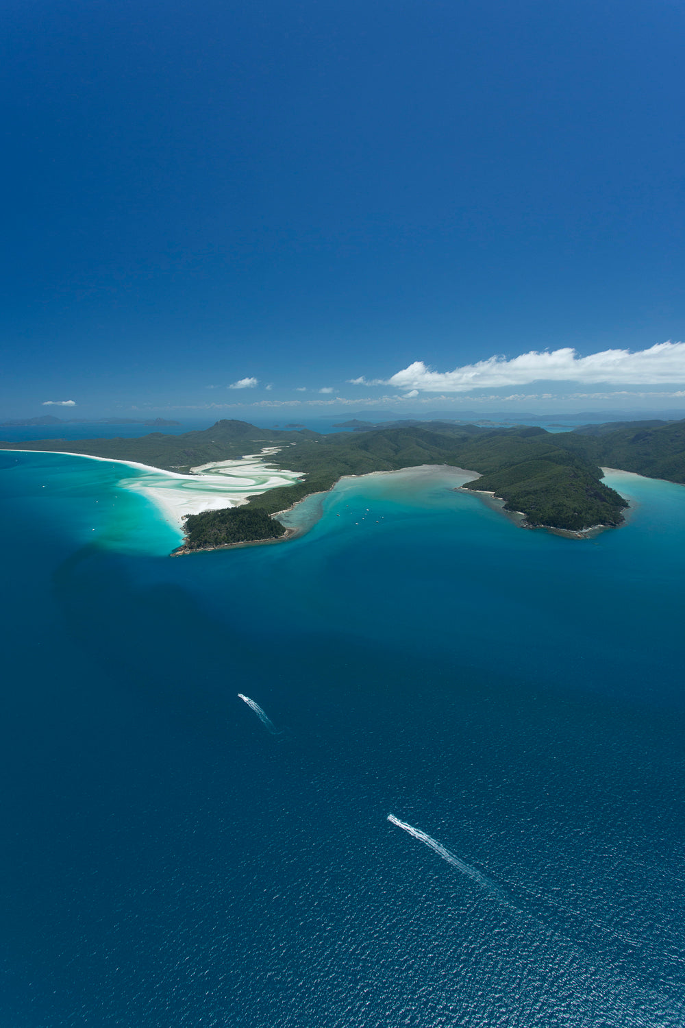 island of green mountains surrounded by white sand swirling in the blue green waters with 2 boats approaching the island