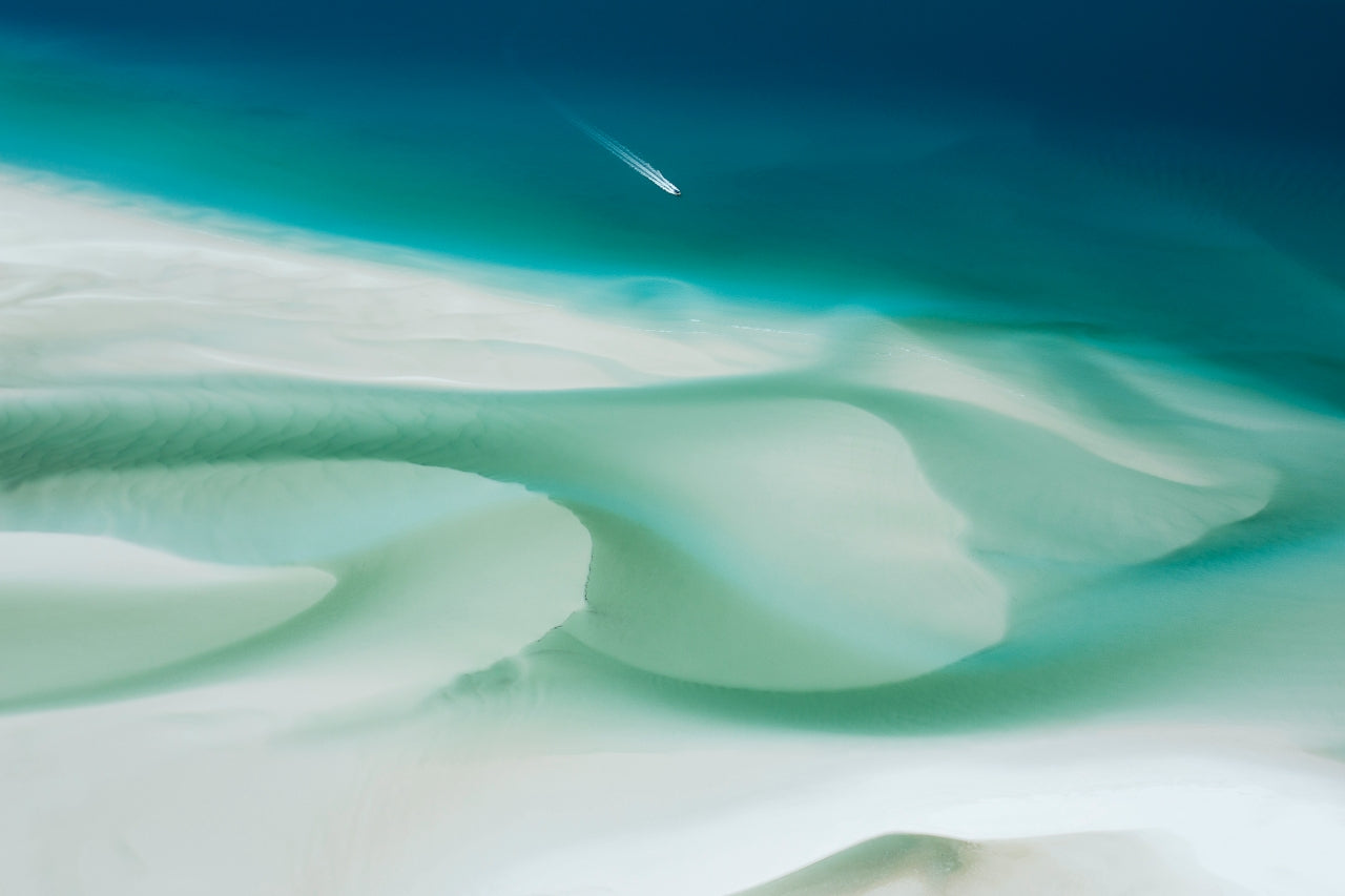 a small boat in the distance replicates a comet amongst the sand and blue green water swirling together