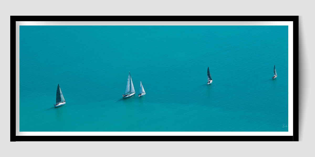 5 sailing vessels with their sails raised in a line, one behind the other with blue ocean surrounding them