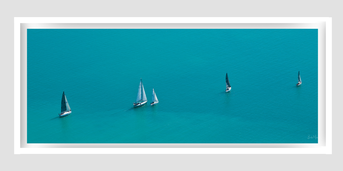 5 sailing vessels with their sails raised in a line, one behind the other with blue ocean surrounding them
