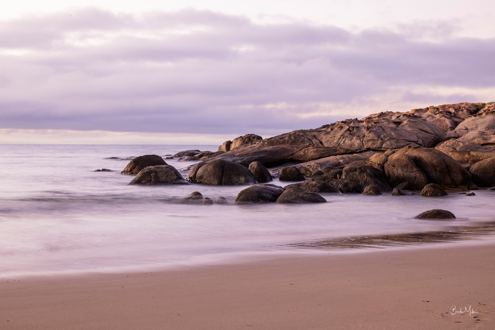 A tranquil beach scene with smooth sand, gentle misty water, large rounded rocks, and a cloudy sky in soft lavender and pink tones.