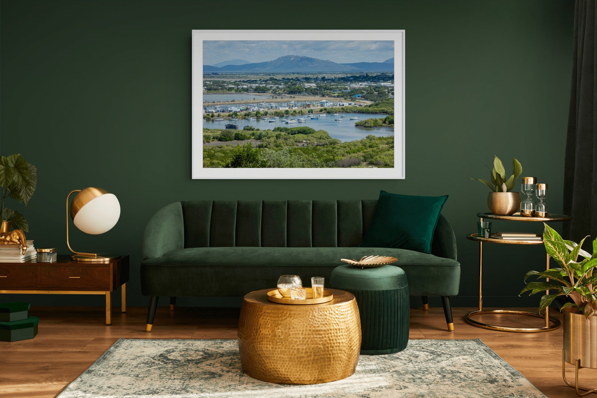 A framed landscape art print of a wetland and river scene hangs on a dark green wall above a matching green velvet sofa in a styled living room.
