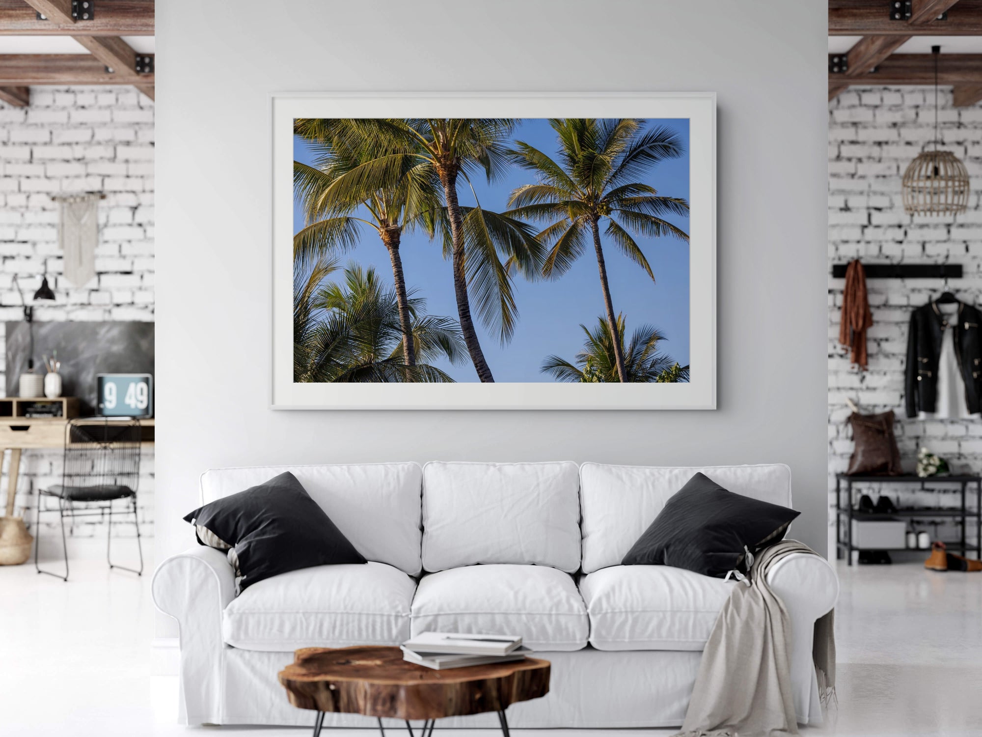 A framed art print depicting tall palm trees against a clear blue sky, displayed on a light gray wall above a white sofa in a modern living room setting.