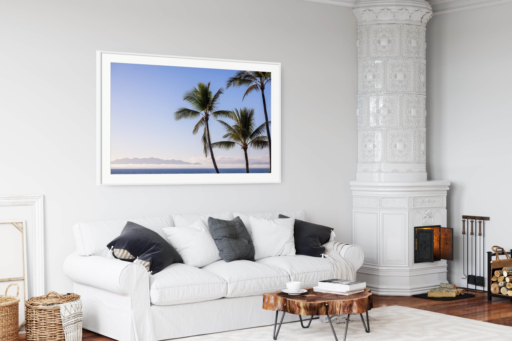 A framed wall art print depicting two palm trees by the ocean at sunset, displayed above a white sofa in a bright, modern living room.