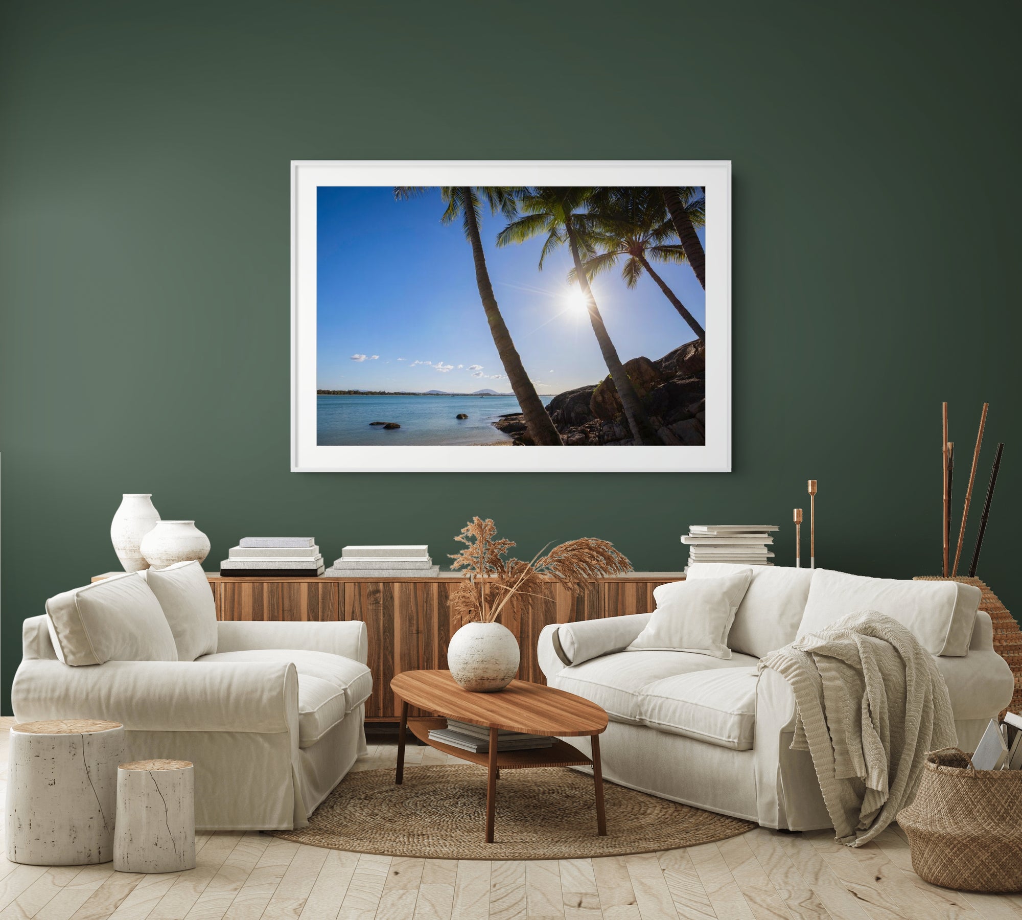A large framed square art print of a tropical beach scene with palm trees and bright sun is displayed on a dark green wall above a styled living room with white sofas, a wooden coffee table, and neutral decor.