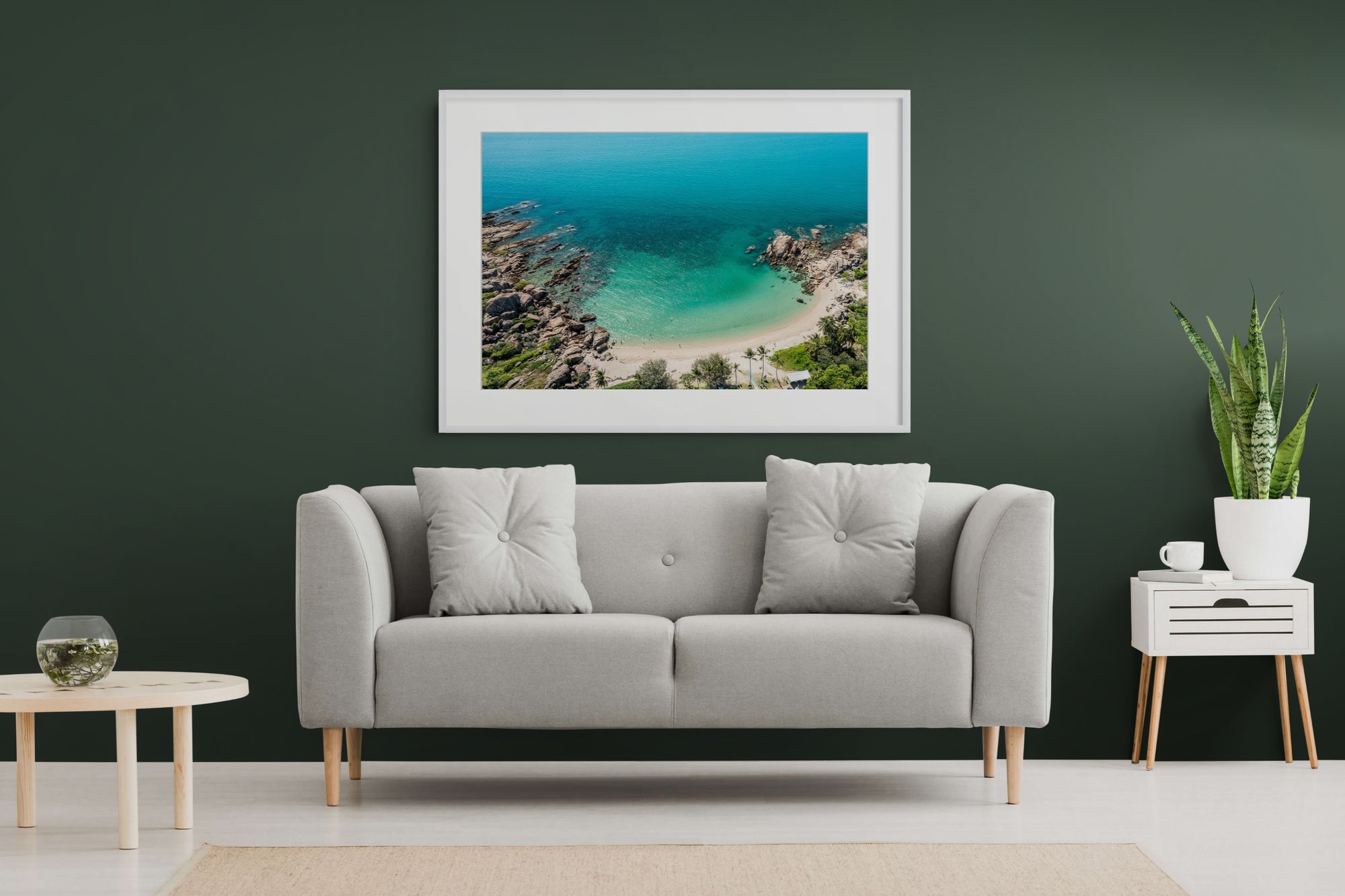 A framed square art print of a tropical beach and turquoise ocean hangs on a dark green wall above a light gray sofa in a styled living room.