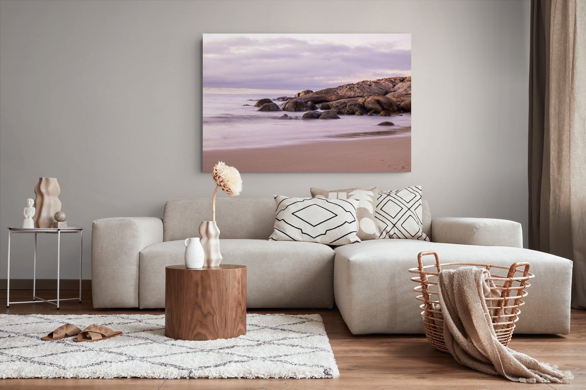A framed wall art print depicting a soft beach and rocky shoreline under a lavender sky, displayed above a light beige sectional sofa in a modern living room setting.