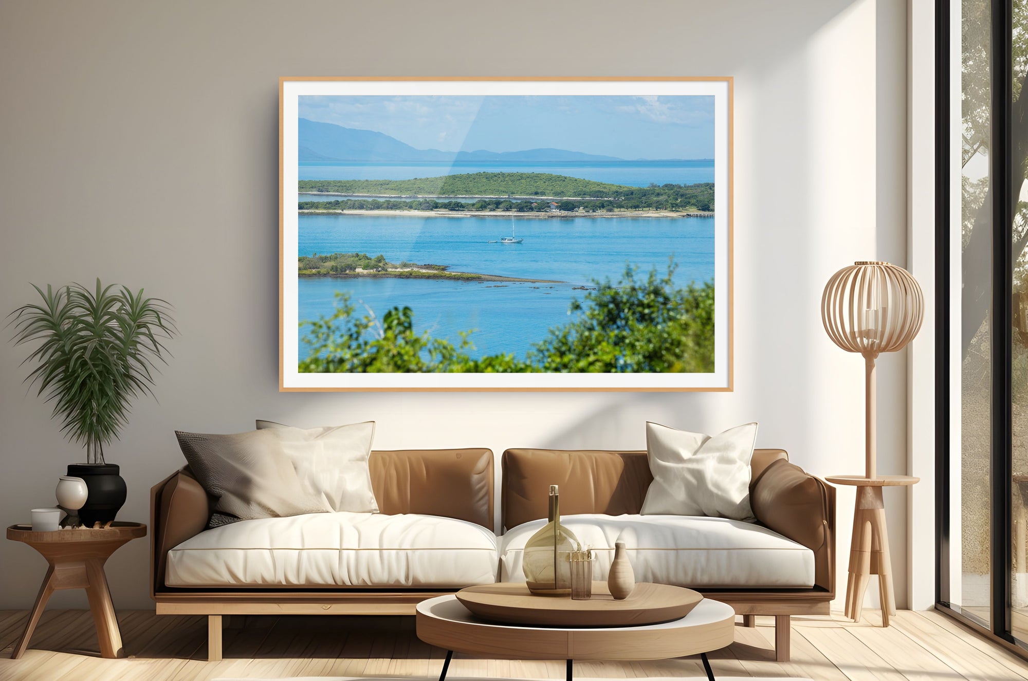 A large framed landscape art print of a coastal bay with blue water, green hills, and distant mountains, displayed on a beige wall above a modern brown and white sofa in a styled living room.