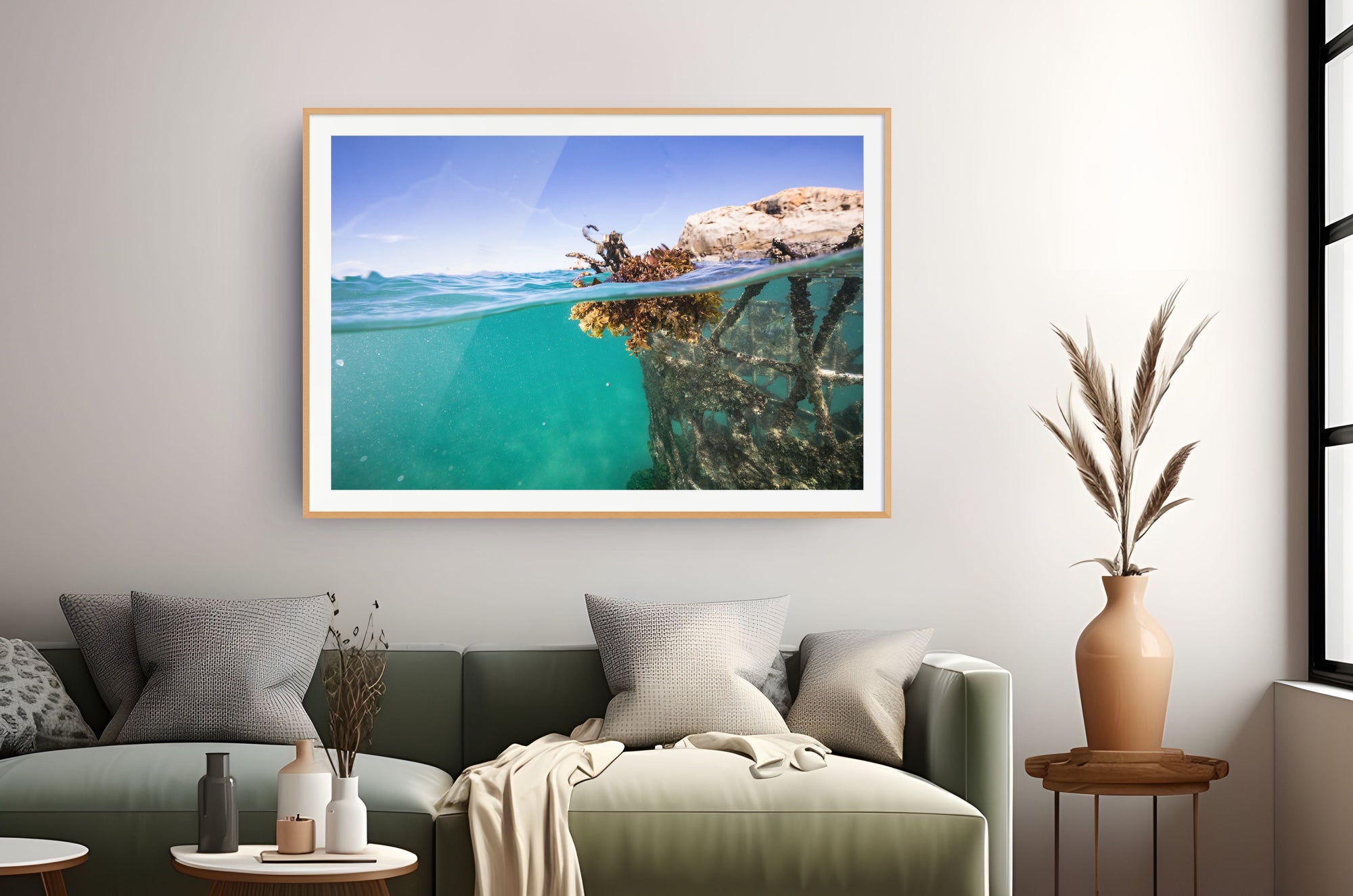 A framed wall art print showing a split underwater and above-water ocean scene hangs above a green sofa in a styled living room. The print depicts turquoise water, rocky shoreline, and blue sky, with a light wood frame and white mat.