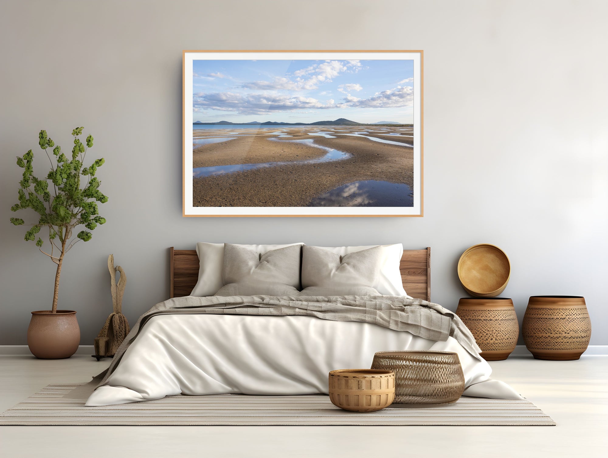A framed landscape art print of a tidal flat or beach with sand, water channels, and distant hills under a cloudy sky, displayed on a light gray wall above a neatly made bed in a modern, minimalist bedroom.