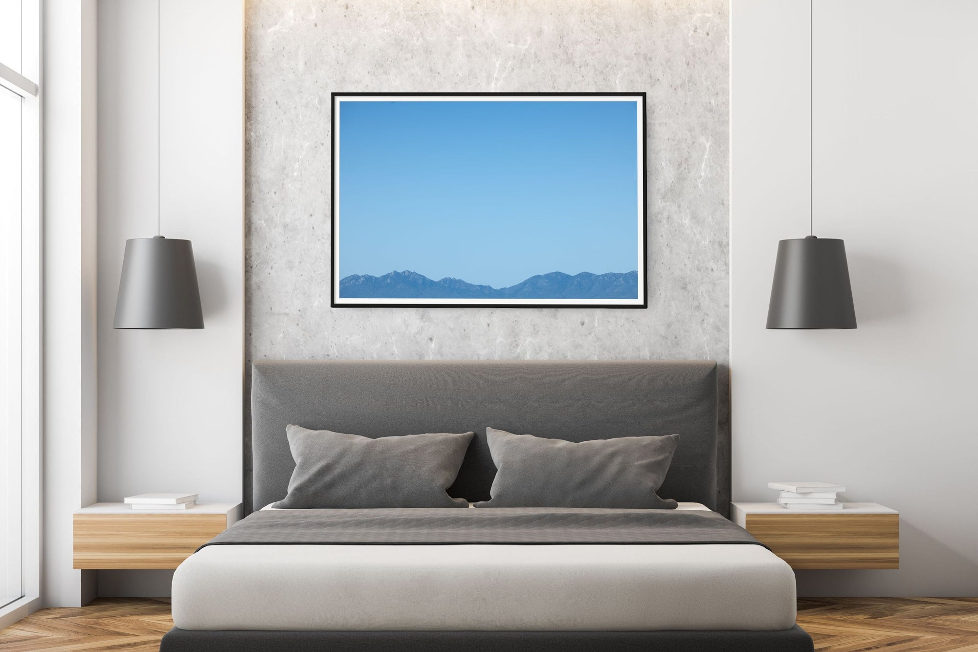 A modern bedroom featuring a low gray platform bed with white and gray bedding, flanked by floating wooden nightstands and black pendant lamps. Above the bed is a textured light gray wall with a framed landscape print showing blue mountains under a clear blue sky.