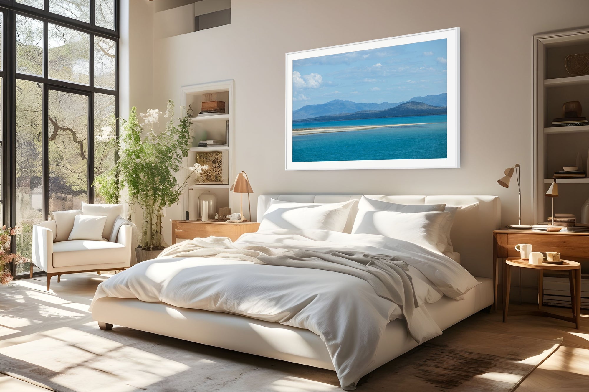 A framed landscape art print of a turquoise sea and mountains hangs on a beige wall above a modern bed in a bright, sunlit bedroom.