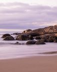 A tranquil beach scene with smooth sand, gentle misty water, large rounded rocks, and a cloudy sky in soft lavender and pink tones.