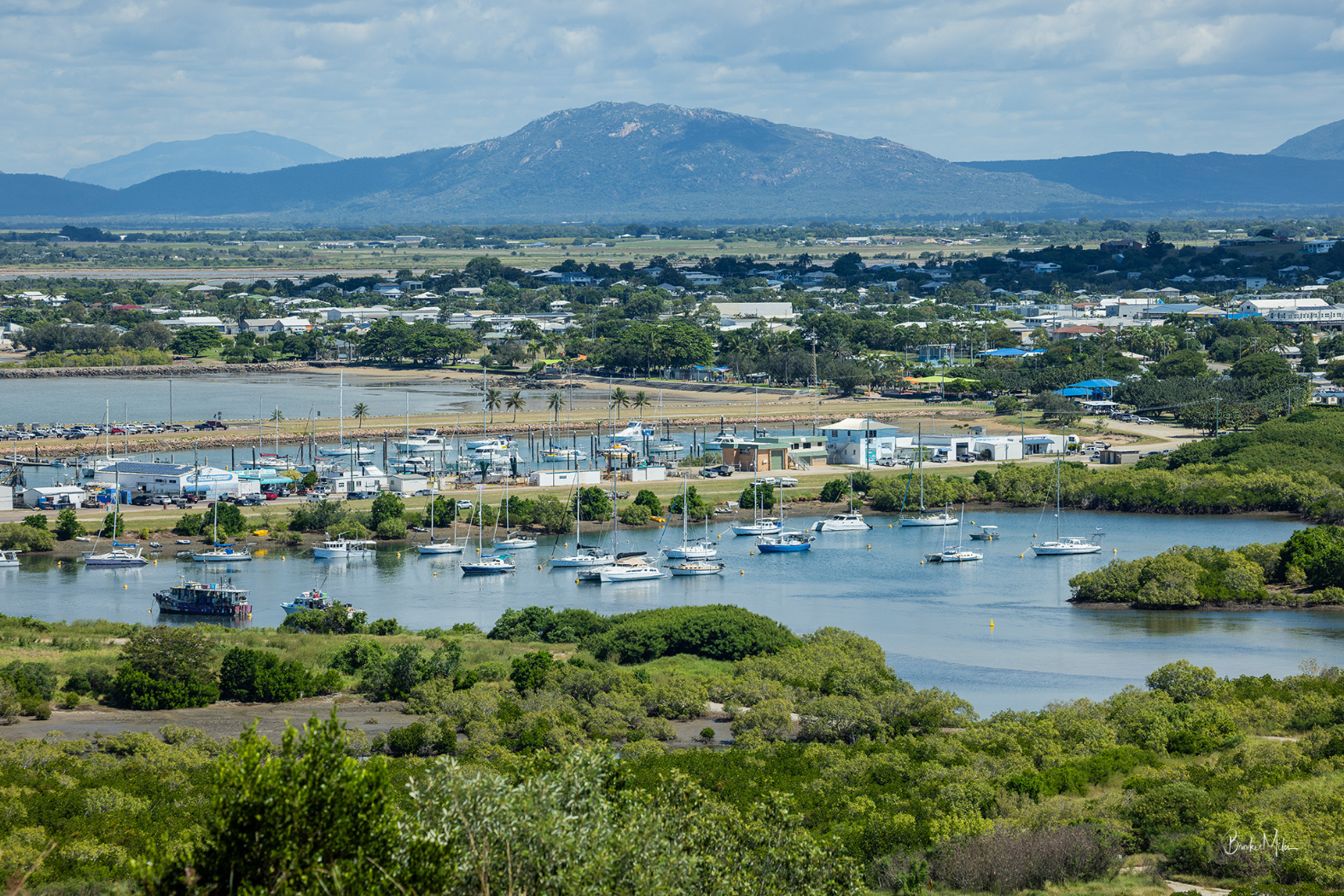 A scenic landscape photograph of a Bowen harbour filled with boats, surrounded by green vegetation and a town, with mountains in the background under a cloudy sky.