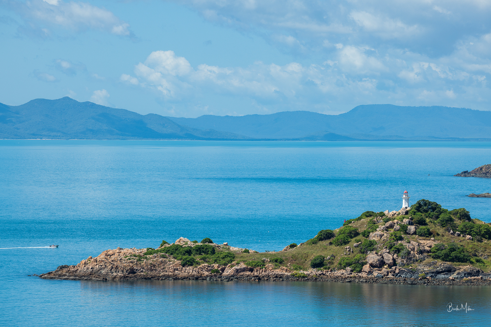 A scenic coastal landscape with bright blue water, distant blue mountains, a rocky island in the foreground, and a small white lighthouse on top of the island under a partly cloudy sky.