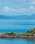A scenic coastal landscape with bright blue water, distant blue mountains, a rocky island in the foreground, and a small white lighthouse on top of the island under a partly cloudy sky.