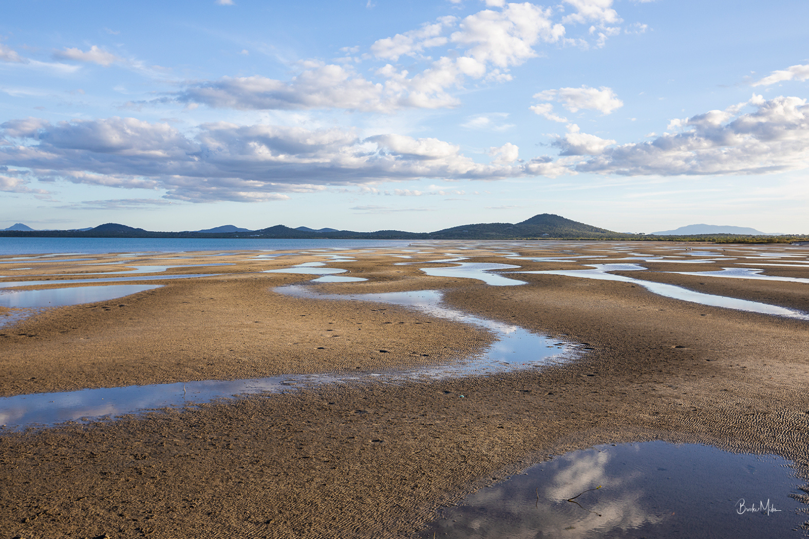 A scenic landscape photograph of a tidal flat or salt marsh with winding shallow water channels reflecting the sky, under a blue sky with scattered white clouds and distant hills.