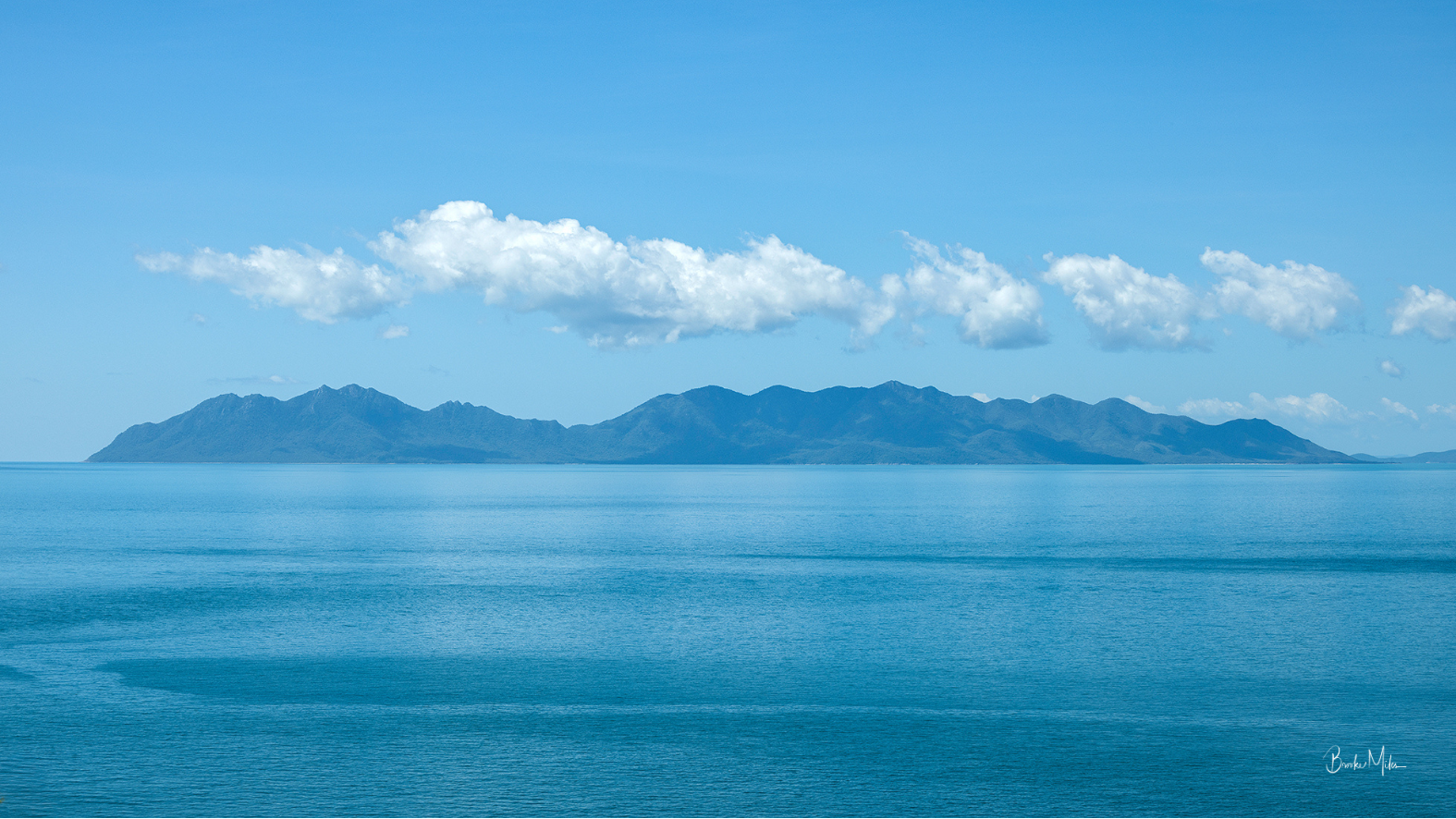 A scenic landscape photograph of a calm blue sea under a bright blue sky with white clouds and distant blue mountains. A small artist signature is visible in the bottom right corner.