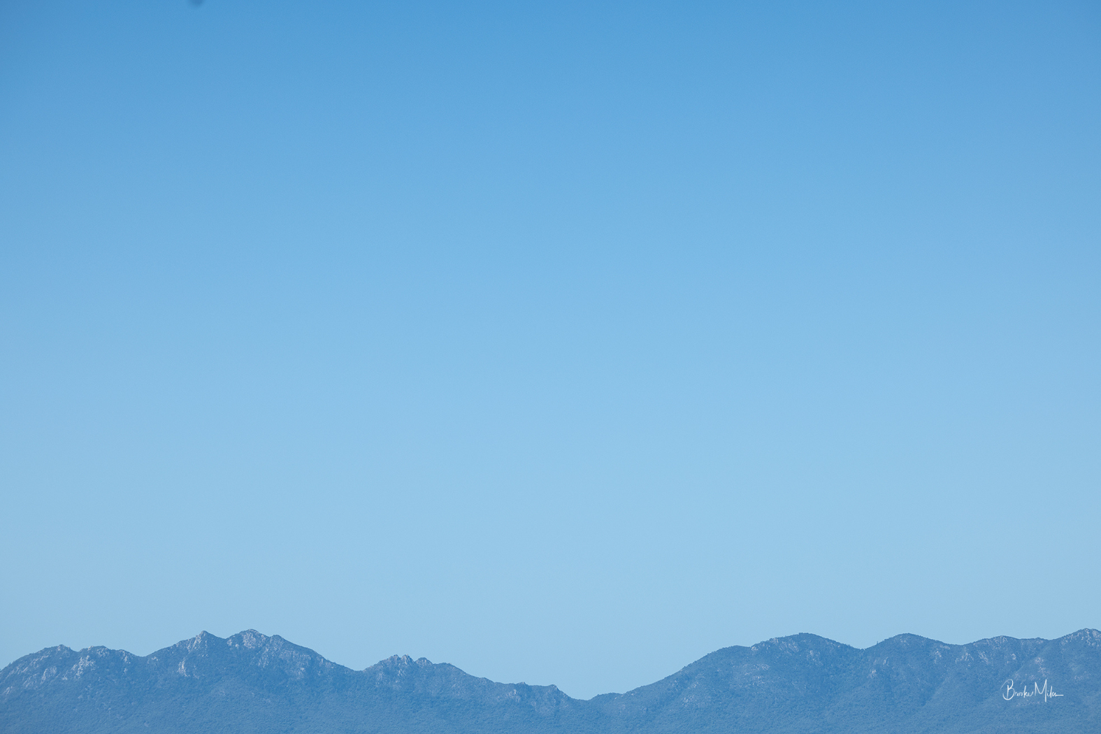 A minimalist landscape photograph featuring a clear blue sky with a distant, hazy mountain range along the bottom horizon.