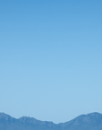 A minimalist landscape photograph featuring a clear blue sky with a distant, hazy mountain range along the bottom horizon.