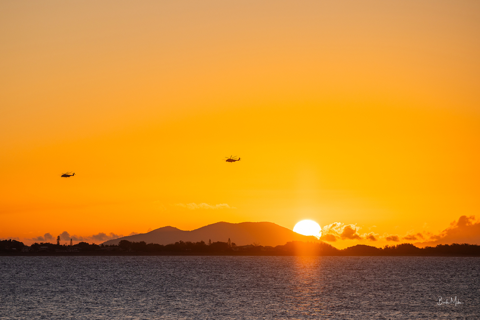 A scenic sunset over water with a mountain silhouette on the horizon, a bright sun near the horizon, and two small helicopters flying across the sky. The sky is a warm golden-orange gradient.