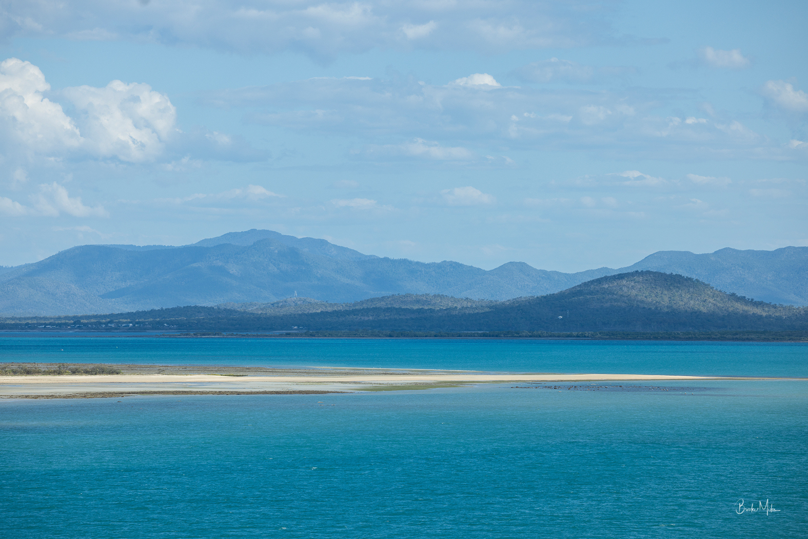 A scenic landscape photograph of a vast turquoise blue lagoon or bay with a narrow sandy spit in the foreground, distant green hills, and blue mountains under a light blue sky with scattered white clouds.