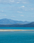 A scenic landscape photograph of a vast turquoise blue lagoon or bay with a narrow sandy spit in the foreground, distant green hills, and blue mountains under a light blue sky with scattered white clouds.