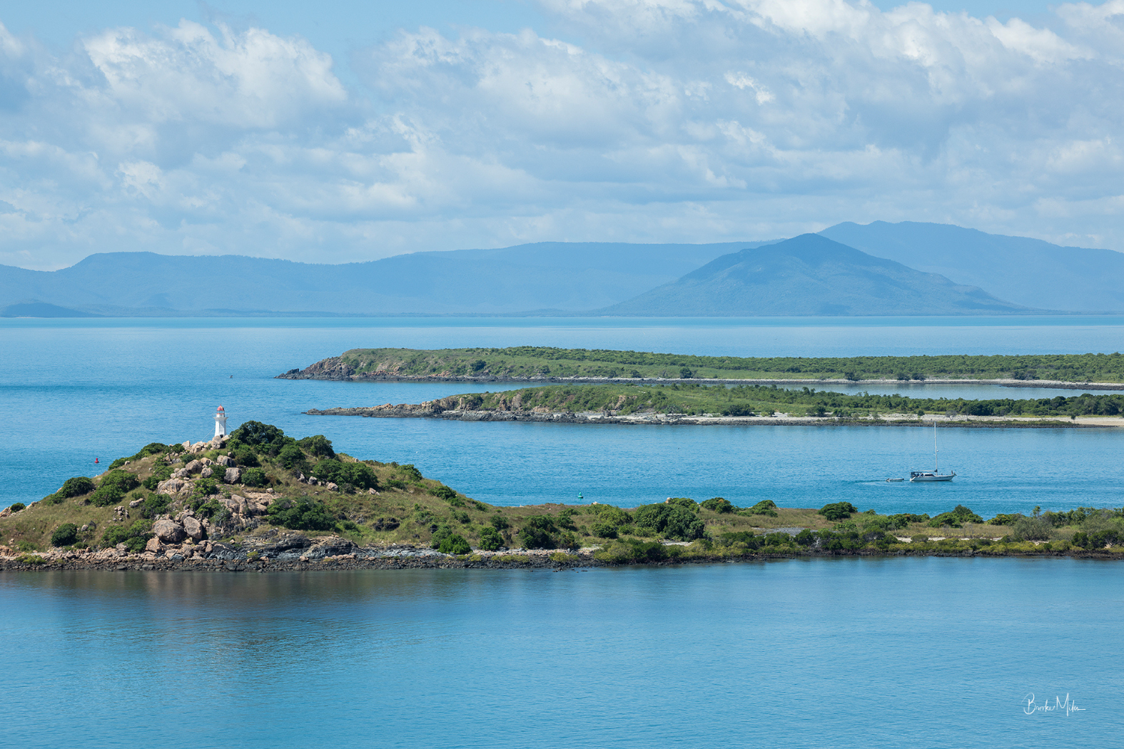 A scenic coastal landscape with bright blue water, small green islands, distant blue mountains, and a cloudy sky. A lighthouse stands on a rocky green island in the foreground.