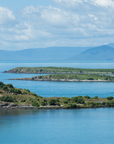 A scenic coastal landscape with bright blue water, small green islands, distant blue mountains, and a cloudy sky. A lighthouse stands on a rocky green island in the foreground.