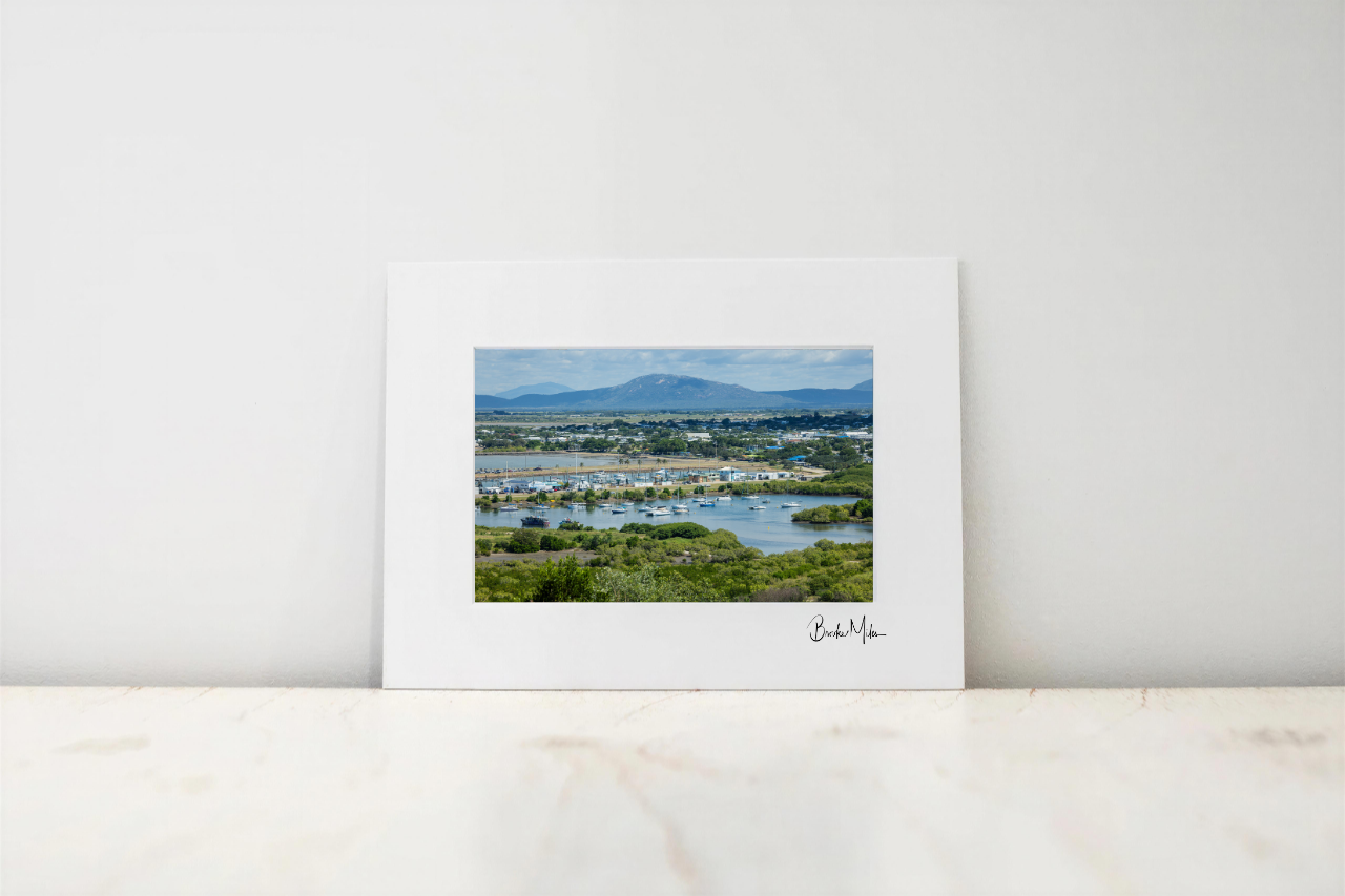A framed landscape art print with a wide white mat border, leaning against a white wall on a light marble surface. The central photograph shows green fields, water, boats, and distant blue mountains.