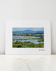 A framed landscape art print with a wide white mat border, leaning against a white wall on a light marble surface. The central photograph shows green fields, water, boats, and distant blue mountains.