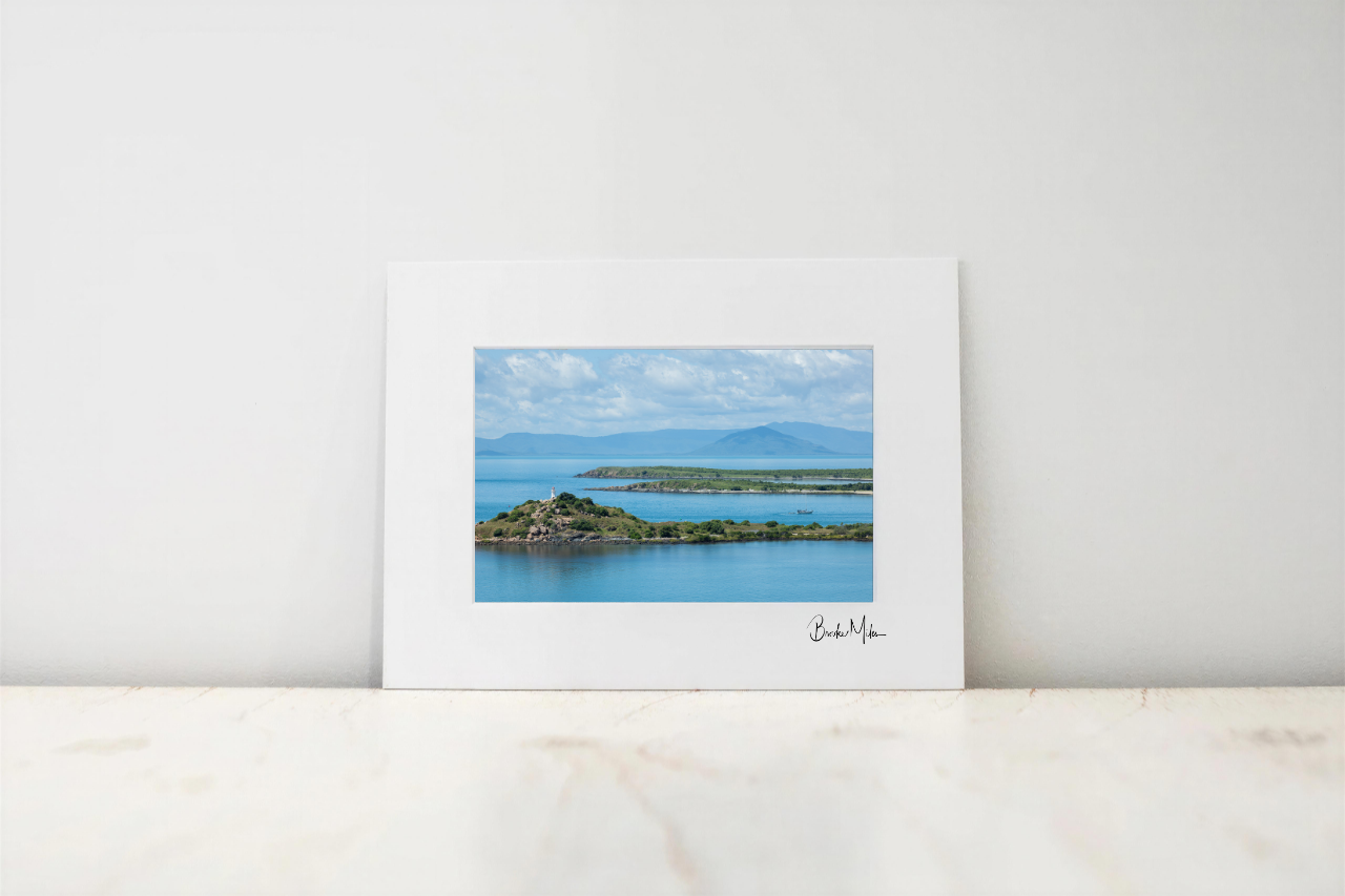 A framed landscape art print featuring a scenic view of a blue bay, green islands, and distant mountains under a cloudy sky, displayed on a light marble surface against a white wall.