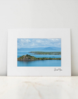 A framed landscape art print featuring a scenic view of a blue bay, green islands, and distant mountains under a cloudy sky, displayed on a light marble surface against a white wall.