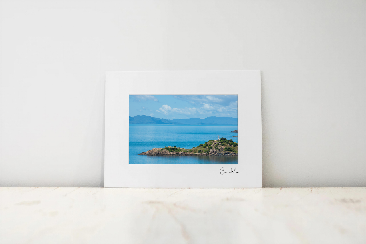 A framed coastal landscape art print with a wide white mat, leaning against a white wall on a light marble surface. The print depicts a blue sea, distant mountains, and a rocky green shoreline under a cloudy sky.