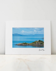 A framed coastal landscape art print with a wide white mat, leaning against a white wall on a light marble surface. The print depicts a blue sea, distant mountains, and a rocky green shoreline under a cloudy sky.
