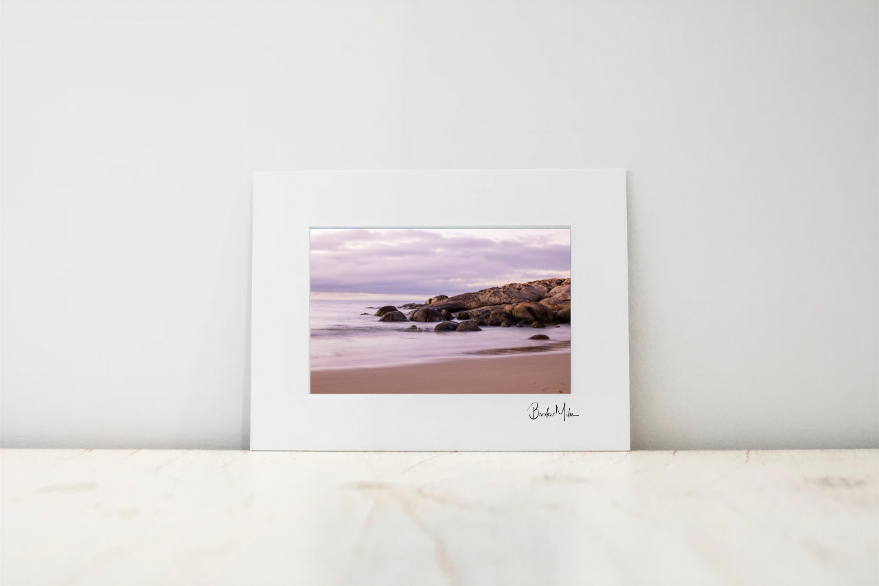 A framed landscape photograph print leaning against a white wall on a light marble surface. The print features a beach scene with rocks, calm water, and a cloudy sky in muted pink, purple, and brown tones, with a white mat border and a small signature at the bottom right.