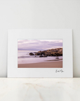 A framed landscape photograph print leaning against a white wall on a light marble surface. The print features a beach scene with rocks, calm water, and a cloudy sky in muted pink, purple, and brown tones, with a white mat border and a small signature at the bottom right.
