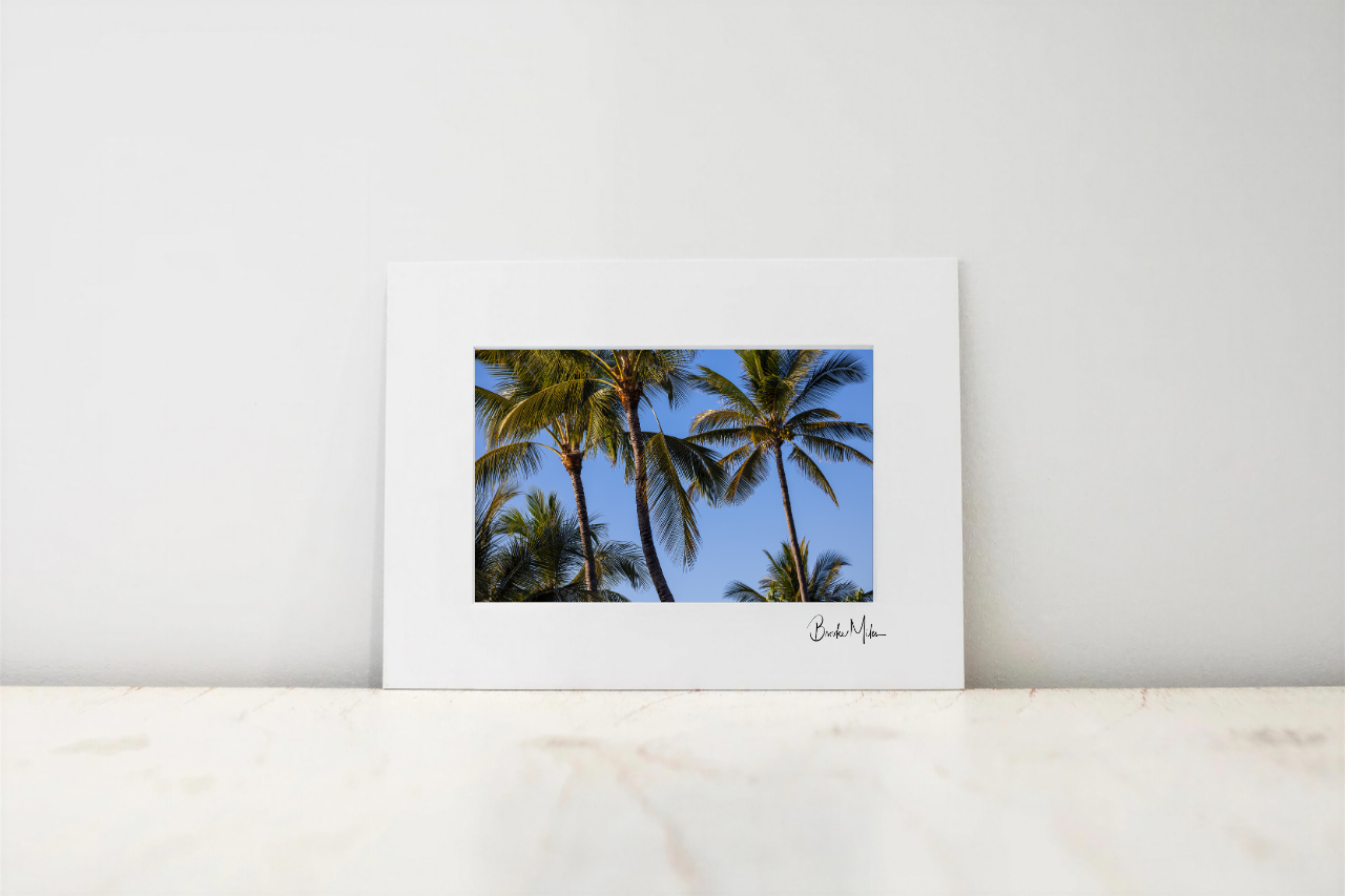 A framed art print featuring a photograph of palm trees against a blue sky, displayed on a light marble surface against a white wall.