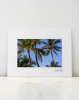 A framed art print featuring a photograph of palm trees against a blue sky, displayed on a light marble surface against a white wall.